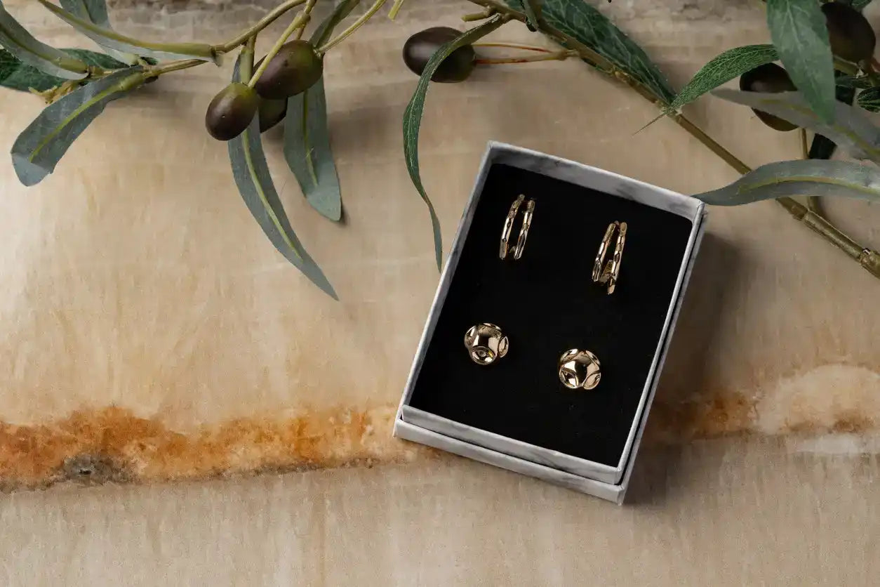 Set of gold hoop earrings in a box on a wooden surface with green leaves.